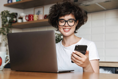 一位戴着眼镜早上拿着手机在家用手提电脑工作的快乐年轻女子的画像