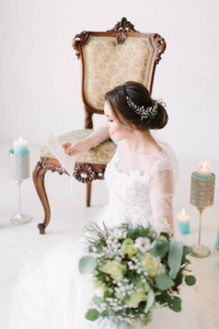 美丽的新娘穿着白色婚纱，坐在椅子附近，蜡烛在窗前的地板上，拿着花束。