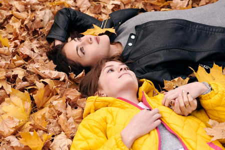 漂亮的女人和十几岁的女孩在秋天公园摆姿势。 他们躺在落叶上。 秋天的美丽景观。