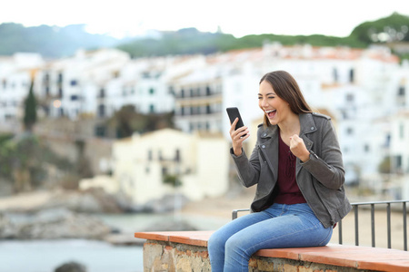 兴奋的女人在度假时坐在窗台上查看智能手机内容