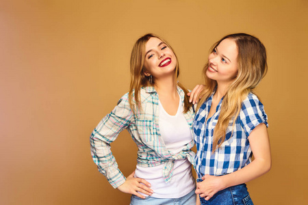 两个年轻漂亮的金发女孩微笑的时髦女孩摆着时髦的夏季格子衬衫衣服。 无忧无虑的女人孤立在金色的背景上。 积极的模特疯狂拥抱