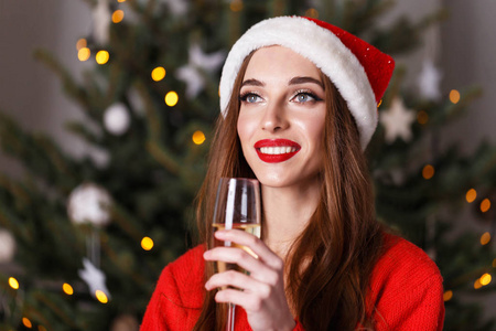 美丽的圣诞妇女穿着圣诞老人帽子喝香槟, 而留在附近的新年云杉