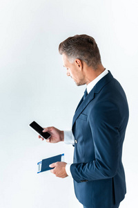 高角度视角的英俊商人使用智能手机与空白屏幕和持有护照与机票隔离在白色