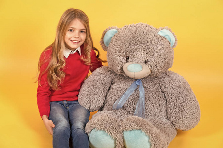 孩子用灰色柔软的玩具微笑。 女孩坐在橙色背景上的大泰迪熊。 带着动物娃娃礼物和礼物的孩子。 节日生日周年庆祝活动。 快乐的童年概