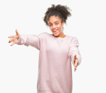 年轻的非洲美国女人穿着冬天的毛衣，在孤立的背景上看着相机，张开双臂微笑着拥抱。 拥抱幸福的快乐表情。
