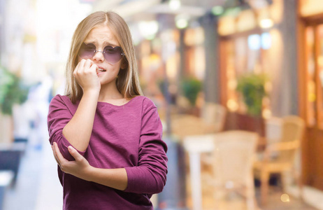 年轻漂亮的女孩戴着太阳镜在孤立的背景上看起来压力和紧张，双手咬着嘴的指甲。 焦虑问题。