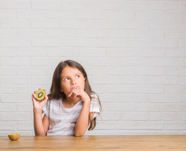 年轻的西班牙裔小孩坐在桌子上吃猕猴桃严肃的脸思考问题非常困惑的想法