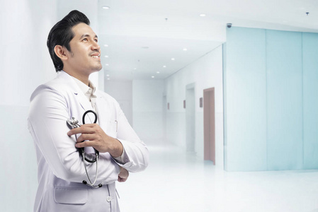友好的亚洲医生男子，白色实验室外套和听诊器交叉手臂站在医院。