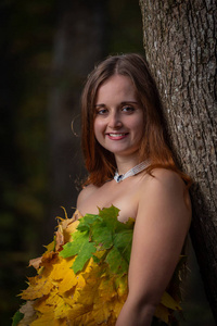 在秋天的森林里，穿着五颜六色的叶子做的连衣裙，一头长发斜倚在树上的美丽的少女