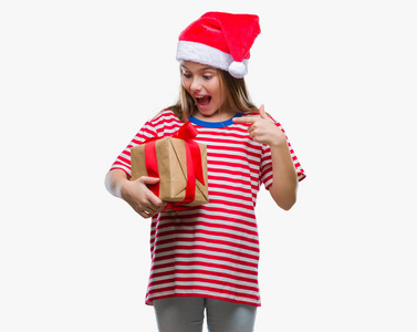 年轻漂亮的女孩戴着圣诞帽，拿着礼物在孤立的背景上，非常高兴地用手和手指指着。