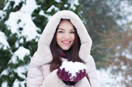 年轻美丽的情感女人的肖像，穿着带帽的羽绒服和手套，在白雪覆盖的花园背景上。 冬季雪景