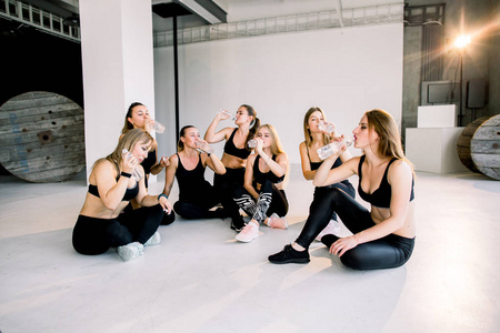 美丽的健身妇女穿着黑色衣服坐在地板上，健身后在健身房喝水