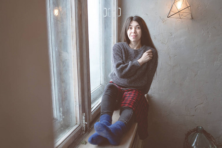 女孩坐在窗户上，一个穿灰色毛衣的学生。 模特对着窗户摆姿势。 概念浪漫与渴望