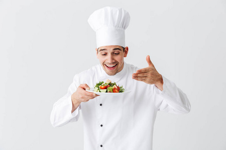 兴奋的男人厨师穿着制服，在白色背景的盘子上展示新鲜的绿色沙拉