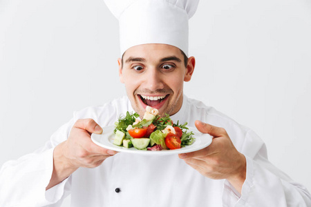 兴奋的男人厨师穿着制服，在白色背景的盘子上展示新鲜的绿色沙拉