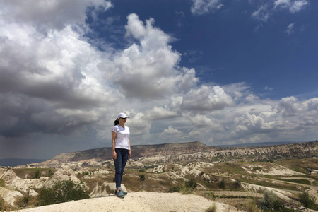 一个穿着白色帽子和T恤蓝色牛仔裤的年轻女孩站在卡帕多西亚的一块岩石上，在多云的天空和山景的背景下眺望远方。
