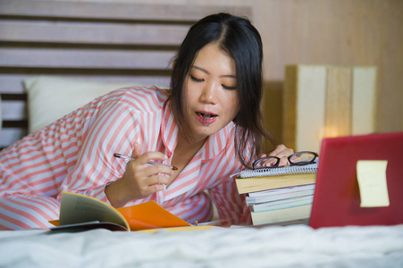 年轻漂亮聪明的亚裔韩国女学生在家，卧室，坐在床上，用笔记本电脑和教科书进行考试，专注于教育概念