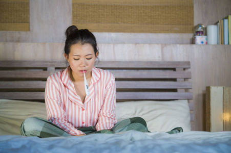 穿着睡衣的年轻美丽甜美的亚裔韩国女孩在家中感到恶心，卧室里患了感冒和流感，体温表在床上覆盖着病毒感染的毯子