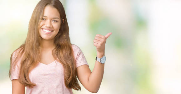 年轻漂亮的黑发女人，穿着粉红色的t恤，在孤立的背景上微笑，快乐的脸看着，用拇指指向一边。