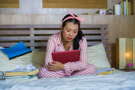 年轻的沮丧和疲惫的亚洲大学生女孩感到不知所措，紧张地准备考试，用笔记本电脑学习，在家里的床上过度劳累