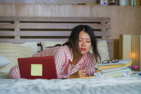 年轻的沮丧和疲惫的亚洲大学生女孩感到不知所措，紧张地准备考试，用笔记本电脑学习，在家里的床上过度劳累