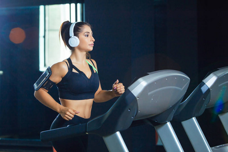 年轻的健康女子在跑步机上跑步, 通过健身房的耳机听音乐。健康生活方式的概念