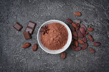白色碗中可可粉的顶部视图，以大块巧克力和整个可可豆为原料，在深灰色混凝土背景下，复制空间