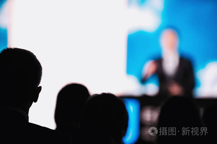 一群商务人士剪影,在会议厅听演讲,模糊的男演讲者穿着西装,天空背景