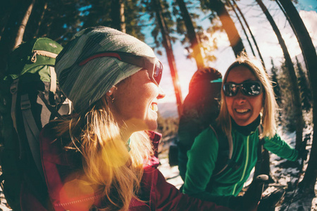 两个微笑的女人在冬季远足。 带着徒步旅行杆的女朋友在白雪覆盖的山路上。 带背包和雪鞋的女孩一起旅行。 朋友们穿过杉树林。