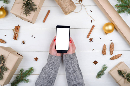 移动智能手机与空白屏幕的应用程序演示在妇女手与装饰复制空间。 木制背景上的圣诞礼物。 平躺顶部视图。 圣诞应用模拟模板