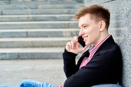 微笑男学生的肖像，独自坐在灰色的石墙附近，在手机上交谈，在大学校园复制空间休息。 教育休息和放松旅行和旅游概念