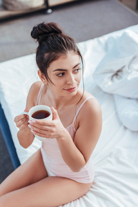 早晨在家中，幸福的年轻女子端着咖啡杯坐在床上