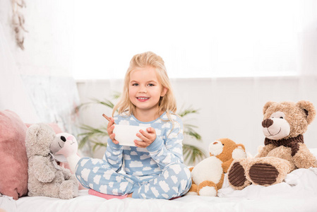 微笑的孩子坐在床上，拿着柔软的玩具，拿着碗吃早餐
