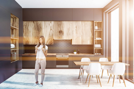 现代厨房内部的女人，灰色的墙壁，混凝土地板，灰色的台面和木制的橱柜，左边是大的灰色和木制的橱柜。有椅子的木制桌子。彩色图像