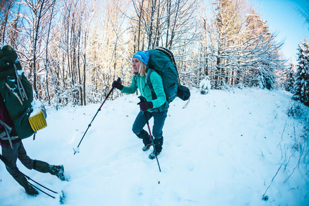 两个女人在冬季远足。 带着徒步旅行杆的女朋友在白雪覆盖的山路上。 带背包和雪鞋的女孩一起旅行。 朋友在阳光明媚的日子穿过杉树林。