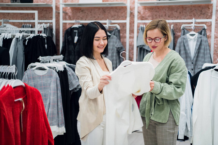 销售顾问帮助在商店为顾客选择衣服。购物与设计师的概念。时尚精品店的女店员和年轻女士