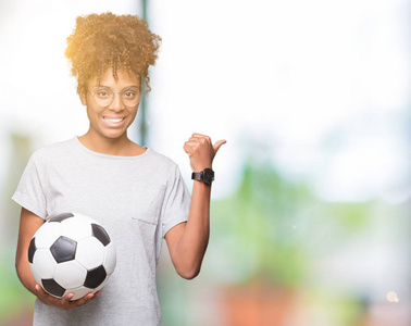 年轻的非洲裔美国妇女手持足球球在孤立的背景上指指点点，并用拇指向旁边露出快乐的脸微笑