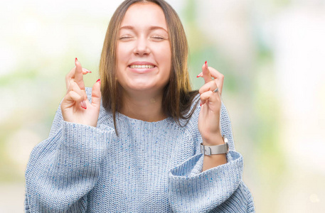 年轻美丽的白种人女人穿着冬天的毛衣，在孤立的背景上微笑着交叉手指，带着希望和眼睛闭上。运气和迷信观念..