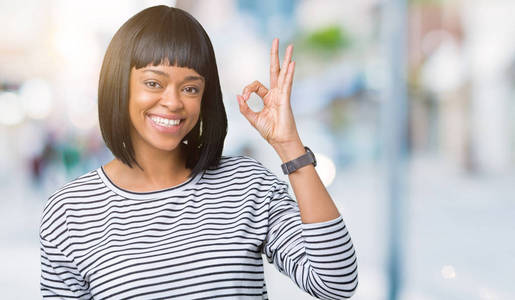 美丽的年轻非洲裔美国妇女穿着条纹毛衣，在孤立的背景上微笑，积极地做好手和手指的标志。 成功的表达。