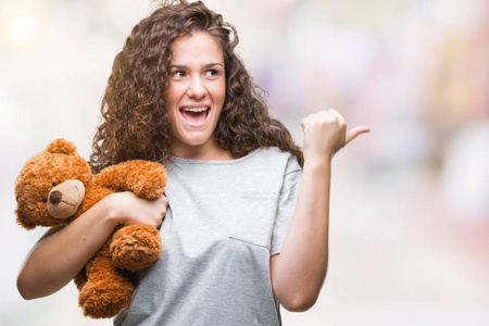 年轻的黑发女孩抱着泰迪熊在孤立的背景上指着，用拇指向侧面展示，脸上带着快乐的微笑