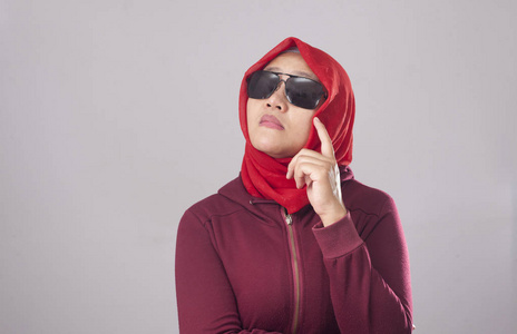 年轻的亚洲穆斯林女士戴着红色西装和头巾的黑色太阳镜的肖像显示出好奇的思维表达。 寻找解决问题的思路