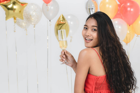 迷人的年轻亚洲女人穿着红色连衣裙，手里拿着饮料，在白色背景上的有趣的派对肖像上，用五颜六色的节日气球快乐。