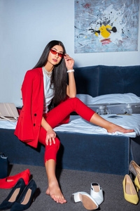美丽的年轻女子穿着时髦的红色西装和太阳镜坐在床上看着相机