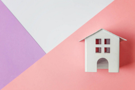 简单的设计与微型白色玩具屋隔离在白色紫罗兰粉红色粉彩彩色时髦的几何背景。 抵押财产保险梦想家居概念。 平躺顶视复印空间