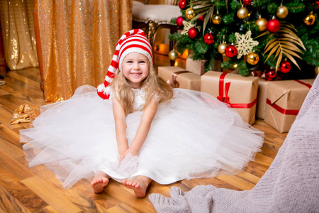 小公主穿着郁郁葱葱的白色礼服和圣诞帽子坐在圣诞树附近。新年快乐和圣诞节主题。节日心情