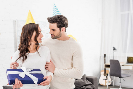 一对漂亮的夫妇戴着派对帽拥抱着，在客厅里拿着生日礼物