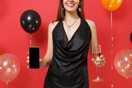 裁剪图像的女孩穿着黑色衣服，拿着一杯香槟手机，空白的黑色空屏在明亮的红色背景气球上。 新年快乐生日模拟节日派对概念