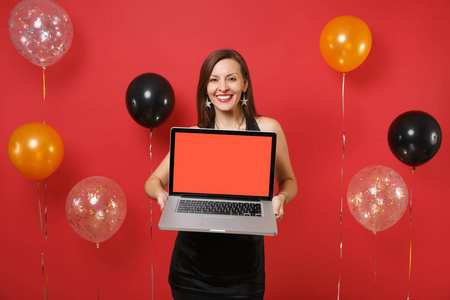 微笑的年轻女子穿着黑色的裙子庆祝，拿着笔记本电脑电脑与空白的黑色空屏上明亮的红色背景气球。新年快乐，生日模拟节日聚会理念