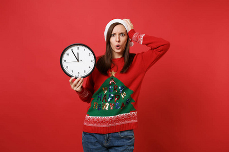 关心的年轻圣诞老人女孩拿着圆钟，把手放在头上，孤立在明亮的红色背景上。 时间不多了。 新年快乐，2019年庆祝节日聚会的理念。 