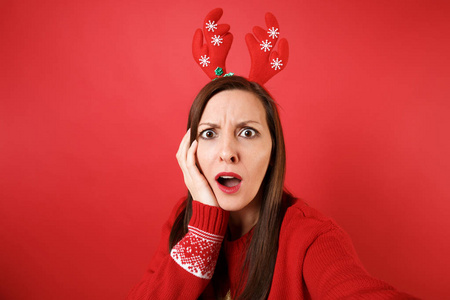 特写自拍的困惑年轻圣诞老人女孩在有趣的装饰鹿角，手放在脸颊上孤立的红色背景。 新年快乐，2019年庆祝节日聚会的理念。 模拟复制
